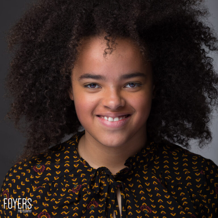 A young person with voluminous curly dark hair smiles at the camera, wearing a patterned black shirt with gold and red designs against a grey background.