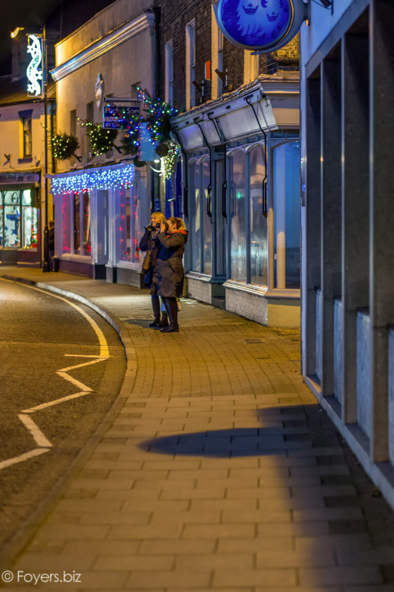A person stands on a nighttime street decorated with festive lights and garlands, photographing the illuminated storefronts.