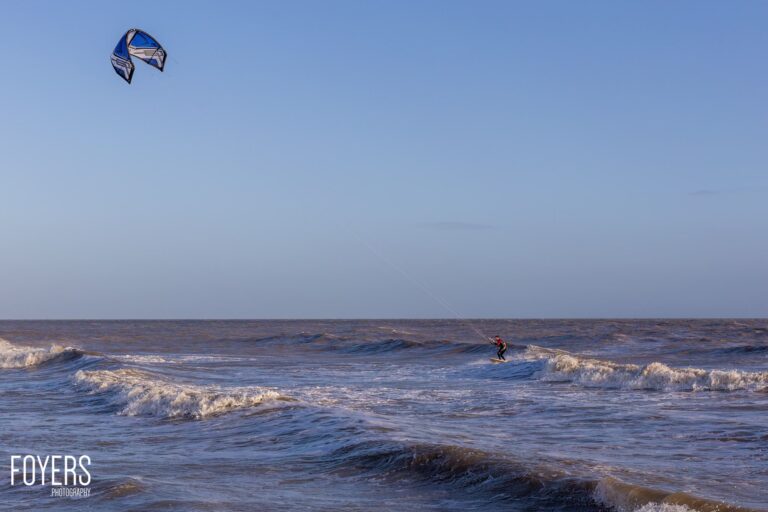 Kitesurfer in the sea with a blue kite above, riding waves under a clear sky.