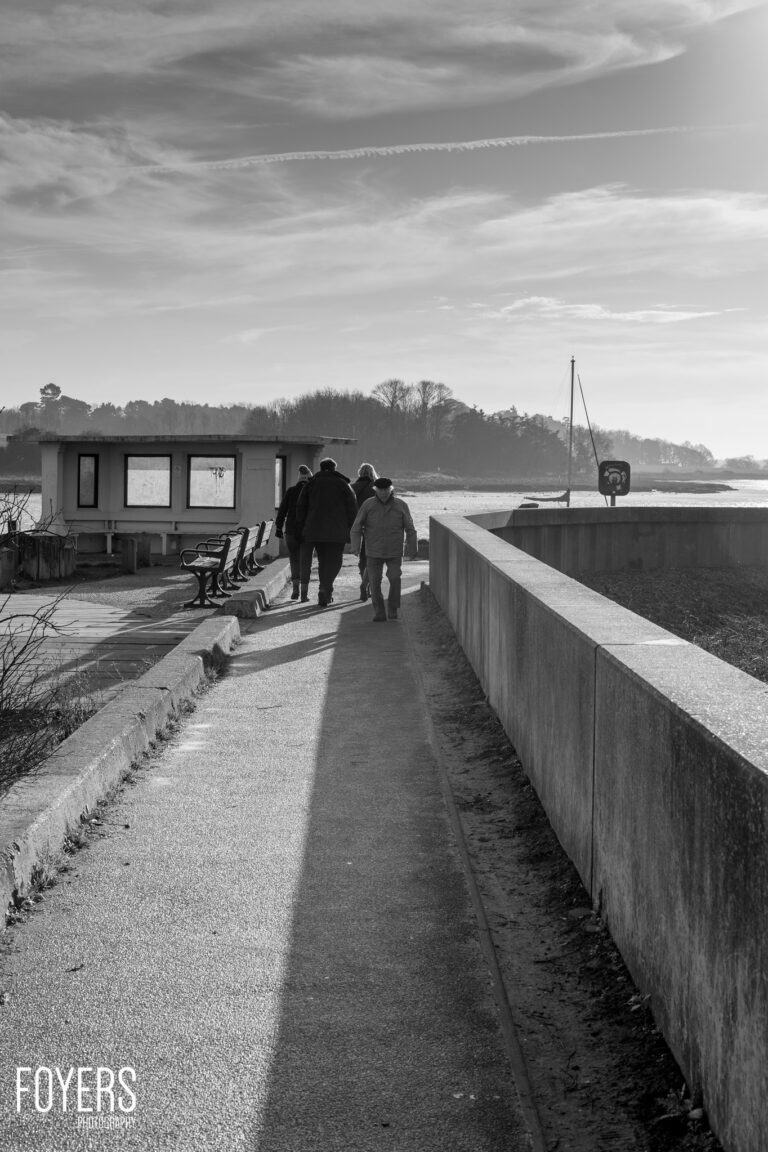 Three people walk along a waterside path past a modern cabin and moored sailboat on a grey winter day.