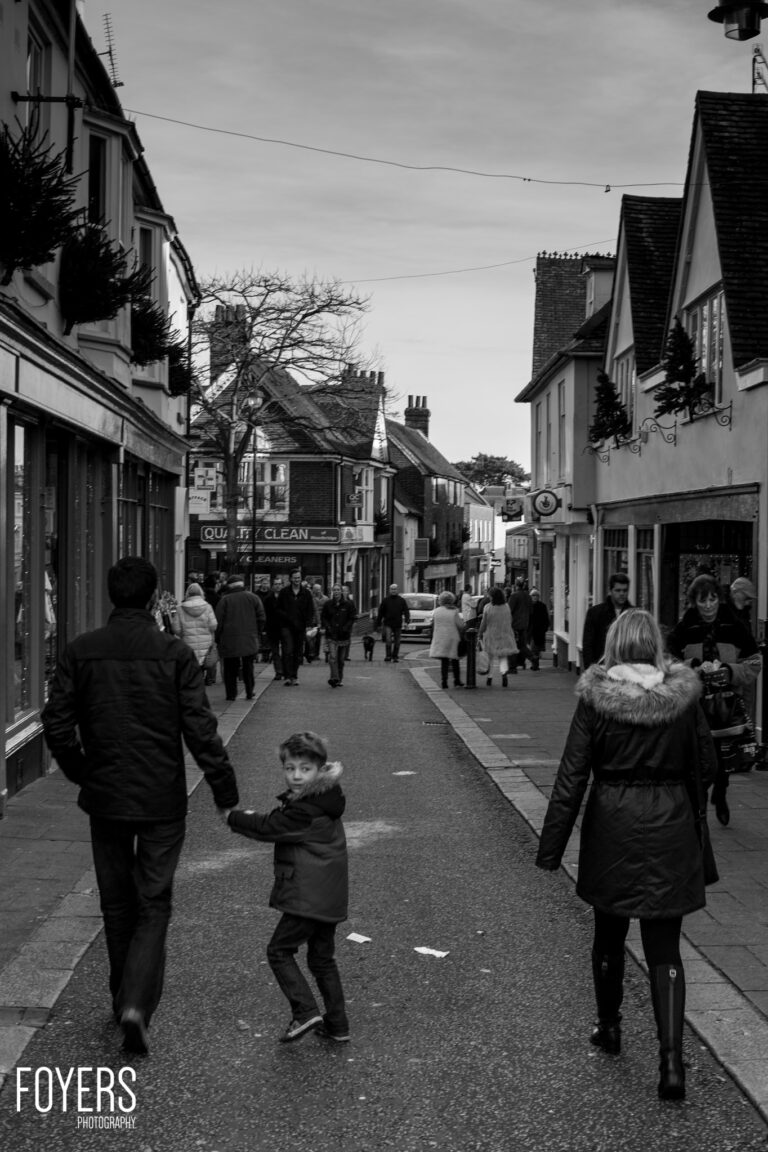 A busy pedestrian high street in black and white. A young child holds an adult's hand while walking among shoppers. Historic buildings line both sides.