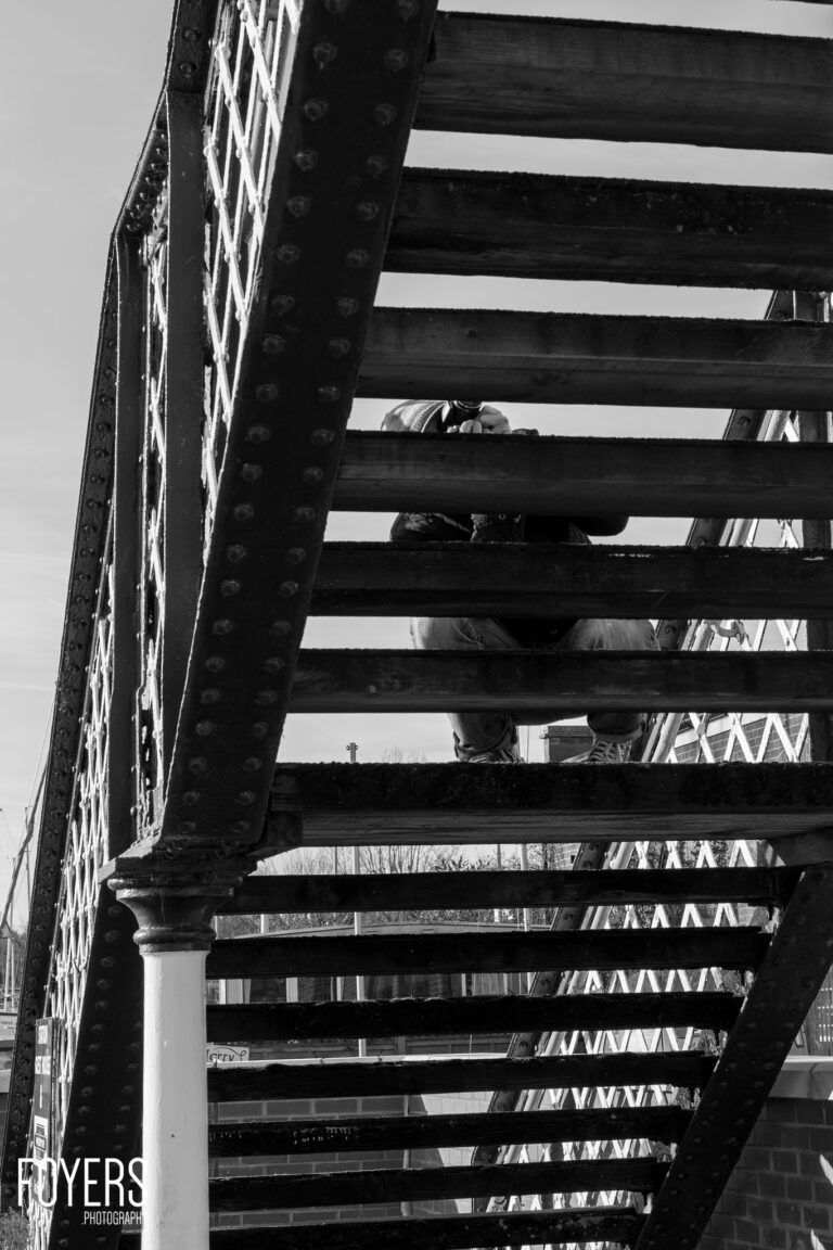 Person sitting on industrial steel beam structure with riveted metal plate on left side, black and white photograph.