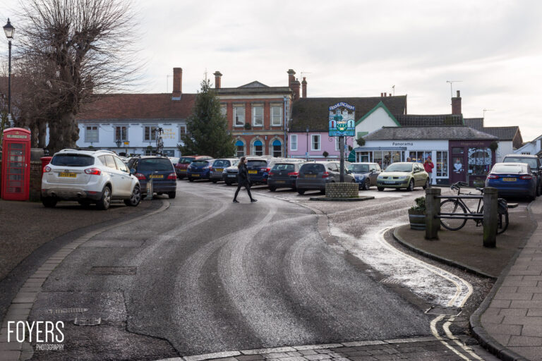 A village high street with parked cars, shops, and historic buildings under an overcast sky.