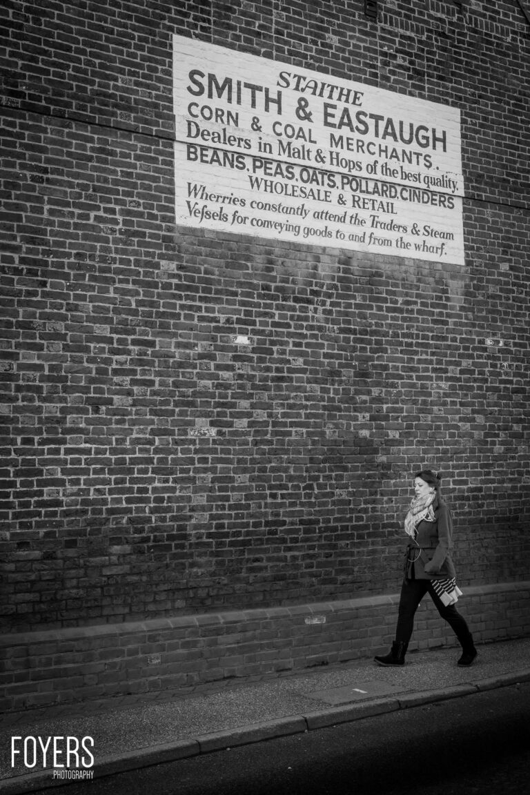 Woman walking past a brick wall with a painted advertisement for Smith & Estaugh, corn and coal merchants.