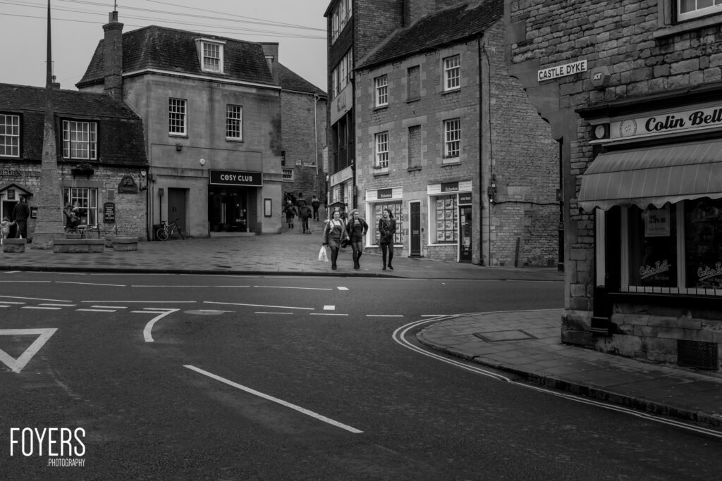 A town square intersection with stone buildings, including a Cosy Club venue and Colin Bell shop. Several pedestrians walk across the street near Castle Dyke.
