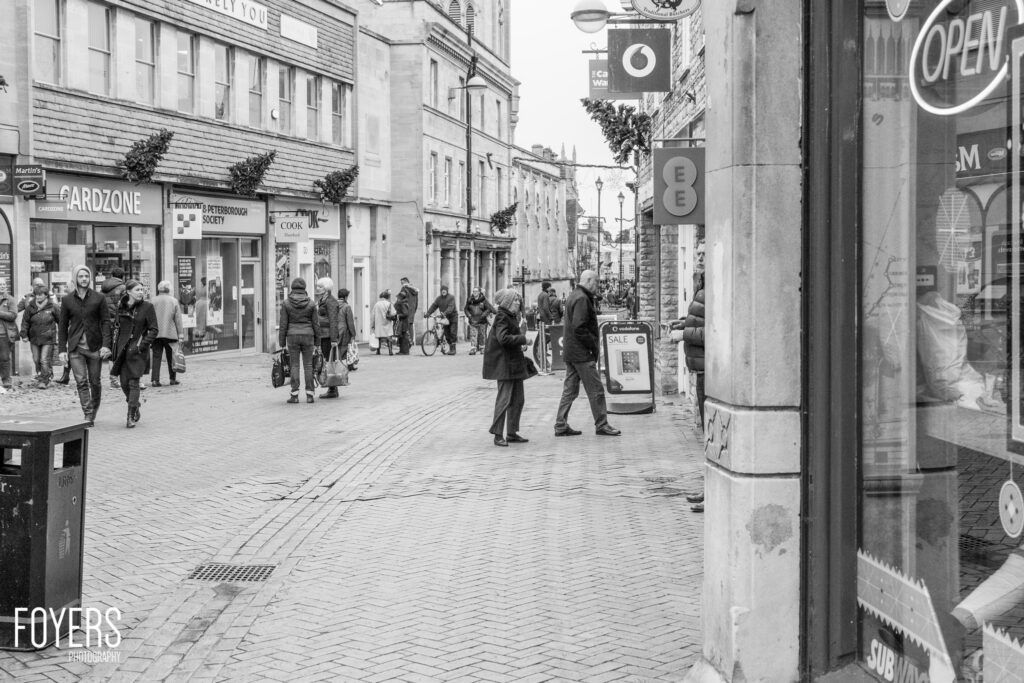 Busy pedestrian shopping street in black and white, with people walking past storefronts including Cardzone and a Peterborough Society shop.