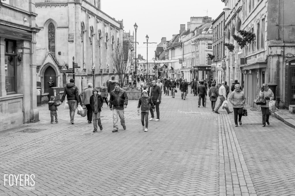 A busy pedestrian street in a historic town centre with Georgian architecture, shopfronts, and a church on the left.