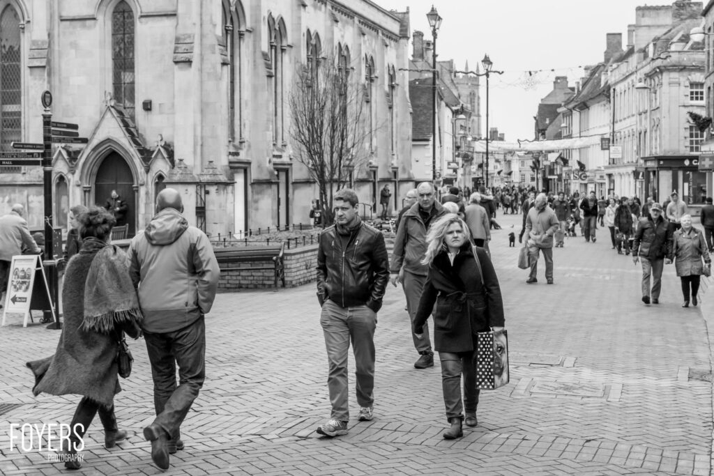 A busy pedestrian street with a Gothic church on the left, Georgian buildings lining both sides, and numerous people walking in winter clothing.