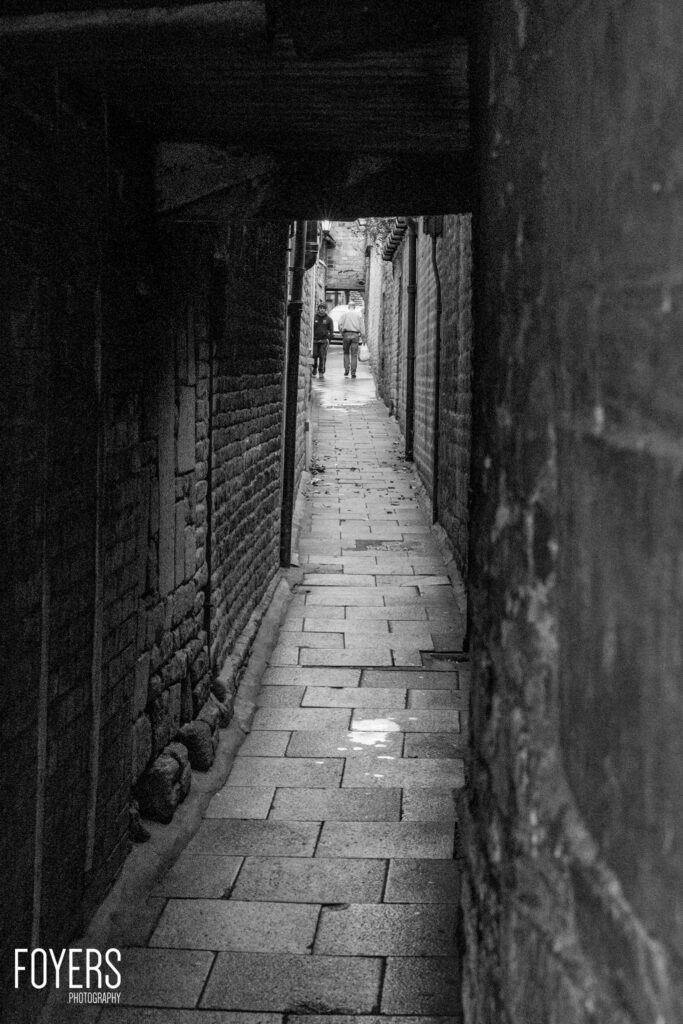 A narrow alleyway between brick buildings with two figures visible in the distance. Black and white photograph.