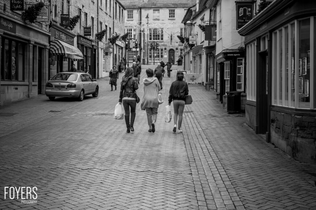 Three women carrying shopping bags walk down a pedestrian high street lined with shops and parked cars, in black and white.