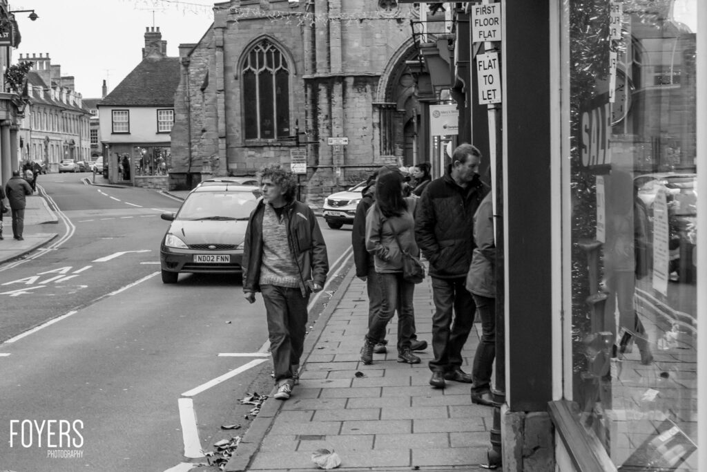 Four pedestrians walking on a pavement past a stone church building in a British high street town center.