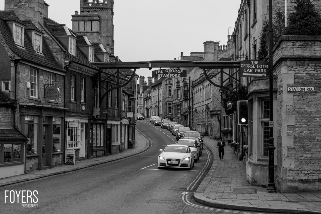 A street scene in Stamford showing parked cars, historic stone buildings, and signage for The George Hotel and Station Road.