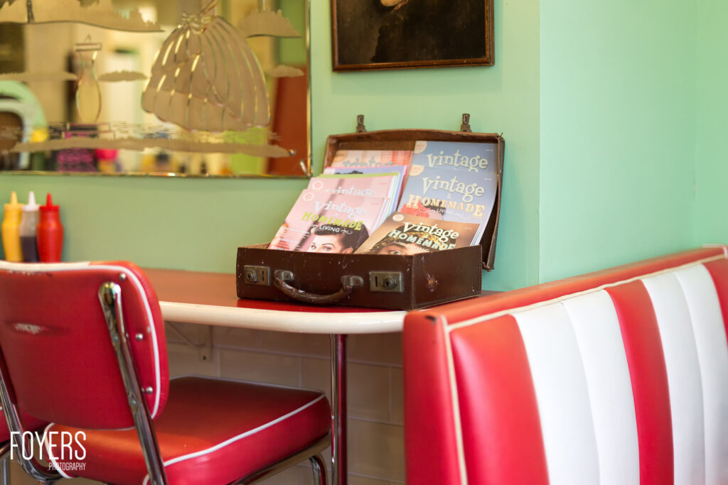 Retro 1950s diner setting with red and white vinyl chairs at a curved counter, vintage suitcase holding magazines, mint green walls.