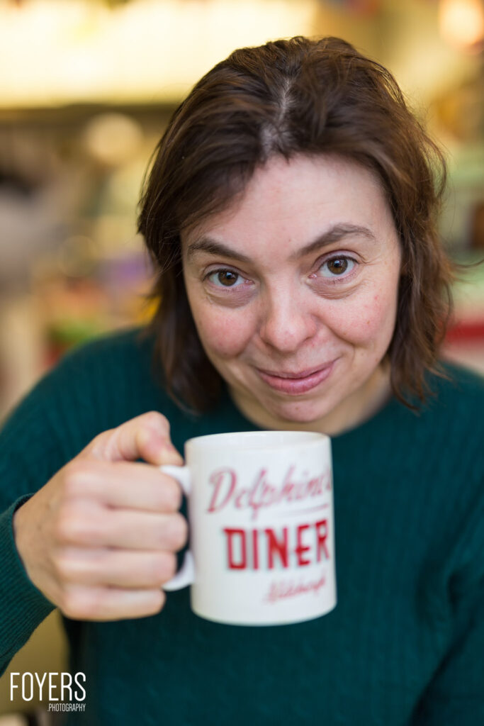 Woman holding a mug reading Dobbin's Diner towards the camera, smiling indoors against a blurred background.