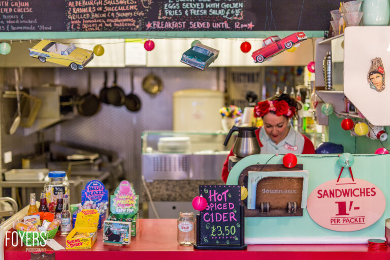A retro-themed diner counter with a staff member wearing red and a flower crown, surrounded by vintage decor, toy cars, and period memorabilia.