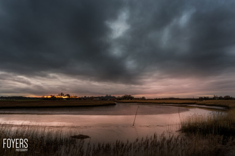 Sunset over a tidal estuary with moody storm clouds, bare reeds in foreground, and distant buildings silhouetted on the horizon.