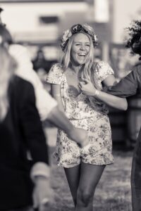 A young woman in a floral dress and flower crown laughs while dancing outdoors at what appears to be a social gathering.