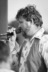 A bearded man with curly dark hair sings into a microphone. He wears a patterned white shirt and has a tattoo on his hand. Black and white photograph.