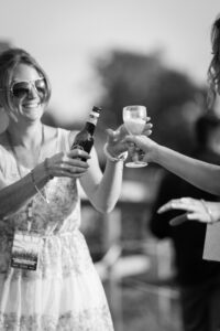 Two women toasting outdoors with a beer bottle and glass, both smiling, in black and white.