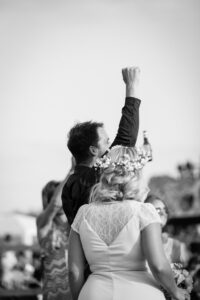A bride and groom celebrate with a raised fist in a black and white wedding photograph. The bride wears a white lace dress and flower crown.
