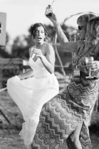 A bride in a white dress and a woman in a patterned dress toast with champagne glasses outdoors at what appears to be a wedding celebration.