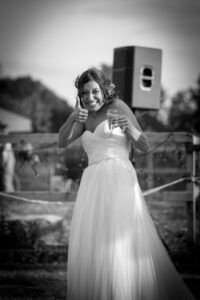 A bride in a white dress gives a thumbs up while holding a champagne flute, standing outdoors with a portable toilet behind her.