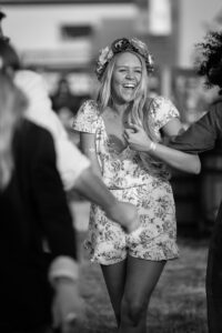 A young woman in a floral dress and flower crown laughs while dancing at an outdoor event, surrounded by other people.