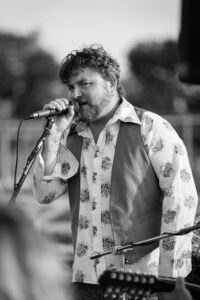 A bearded man in a patterned shirt and waistcoat sings into a microphone at an outdoor performance.