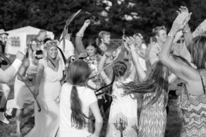 A bride in a white dress and crown dances with guests at an outdoor wedding celebration, arms raised joyfully.