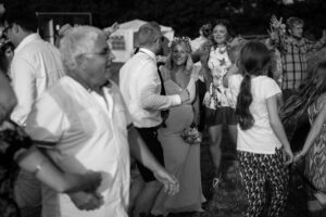 A bride in a tiara dances with guests at an outdoor evening celebration, surrounded by smiling people in casual attire.