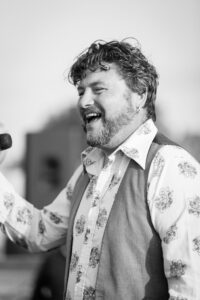 A man with dark curly hair and a beard laughs while singing into a microphone, wearing a patterned shirt over a vest.