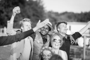 A group of smiling people celebrate outdoors, with one man holding up a camera. Trees line the background.