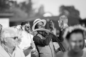 A person in a straw hat and patterned shirt claps their hands while standing among a group of people outdoors in this black and white photograph.