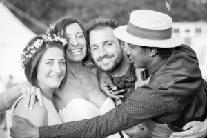 Four people smiling at the camera in a black and white photo. A bride in a white dress and floral crown stands with three others in casual attire outdoors.