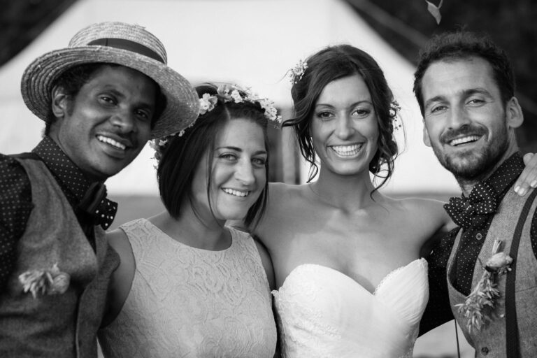 Black and white photo of a bride with three guests at a wedding, all smiling at the camera outdoors.