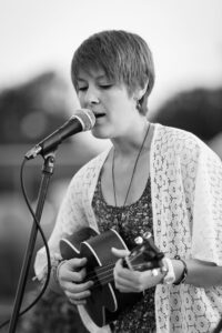 A woman with short hair sings into a microphone while playing an acoustic guitar outdoors in black and white.