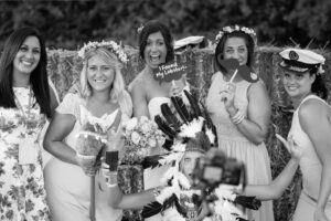 A group of women in white dresses and flower crowns pose together outdoors, holding props and smiling at the camera.