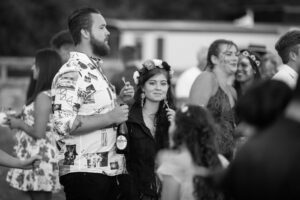 Black and white photo of a bearded man in a patterned shirt standing with a woman wearing a flower crown at an outdoor gathering.