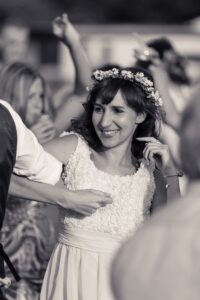 A bride in a white rosette-textured dress and flower crown smiles while being embraced at her wedding celebration.
