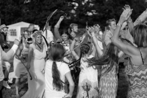 A bride in a white dress and floral crown dances with guests at an outdoor wedding celebration in black and white.
