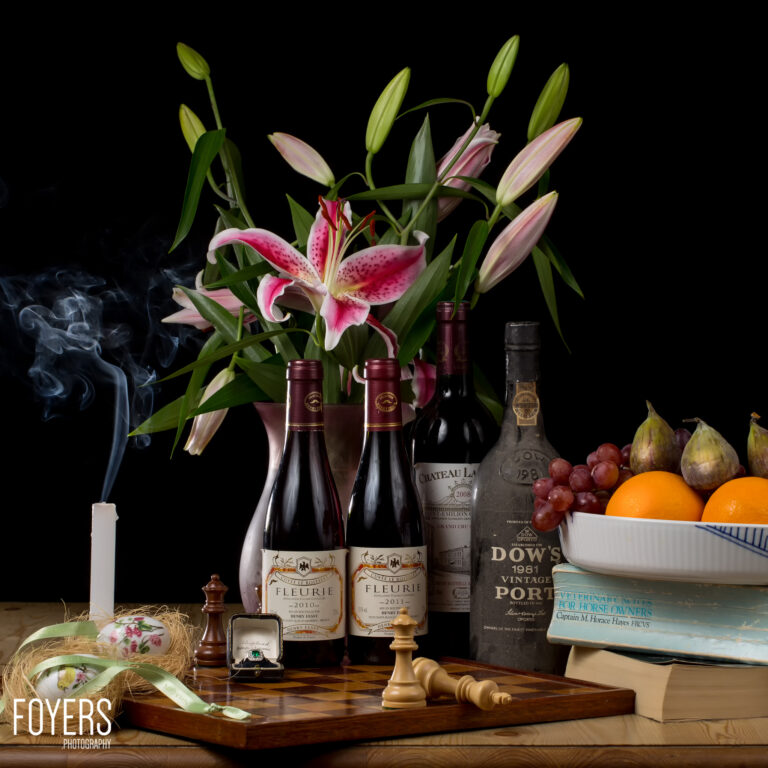 Still life arrangement featuring pink lilies in vases, wine bottles, fresh fruit, books, and decorative objects on a wooden surface against a black background.