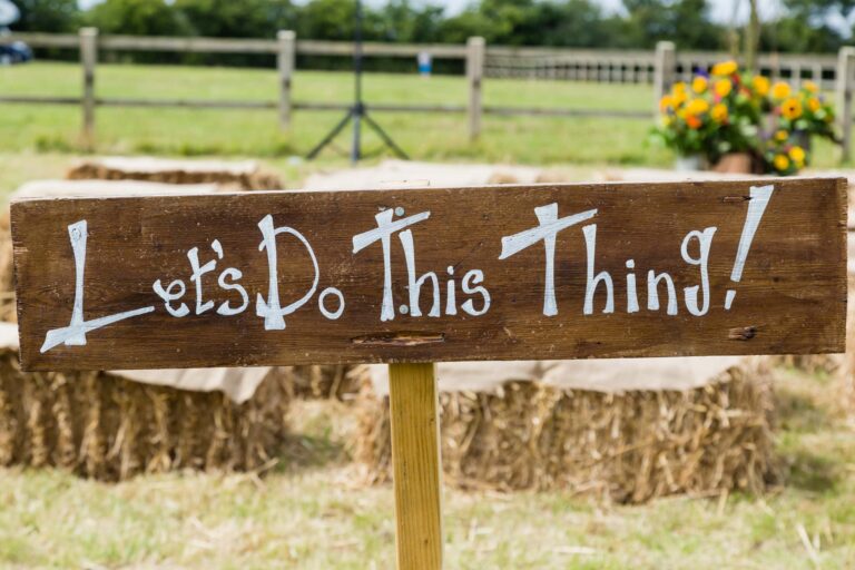 A wooden sign reading Let's Do This Thing! in white paint, mounted on a post at what appears to be an equestrian event venue with fencing and flowers visible behind it.