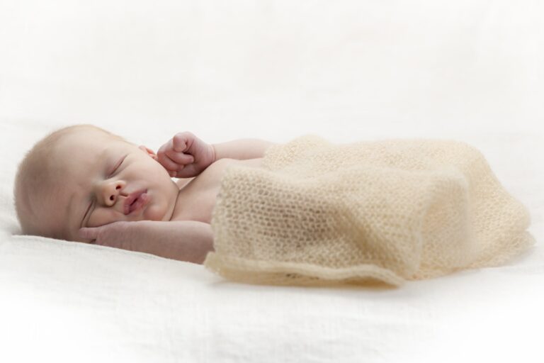 A newborn baby sleeps peacefully on white bedding, swaddled in a cream knitted blanket.