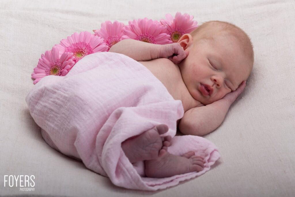 A newborn baby wrapped in pink fabric, sleeping peacefully on a white background with pink gerbera flowers arranged above.