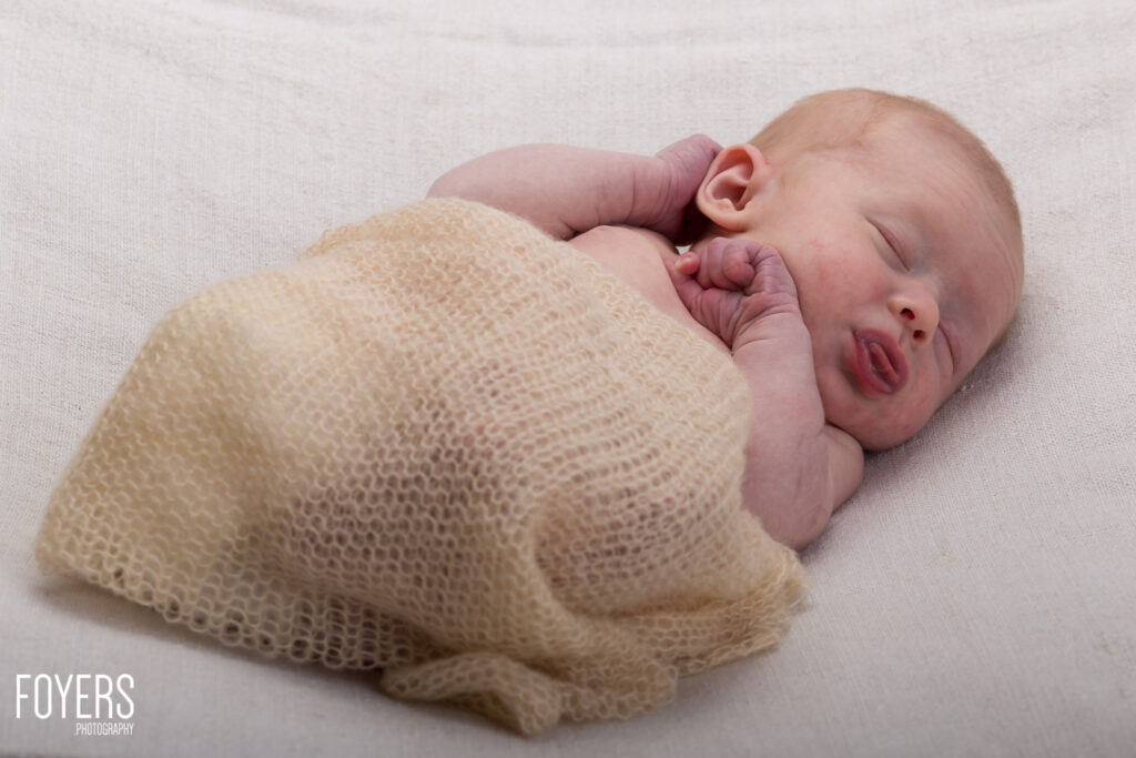 Newborn baby swaddled in cream knitted wrap, sleeping on white fabric background.
