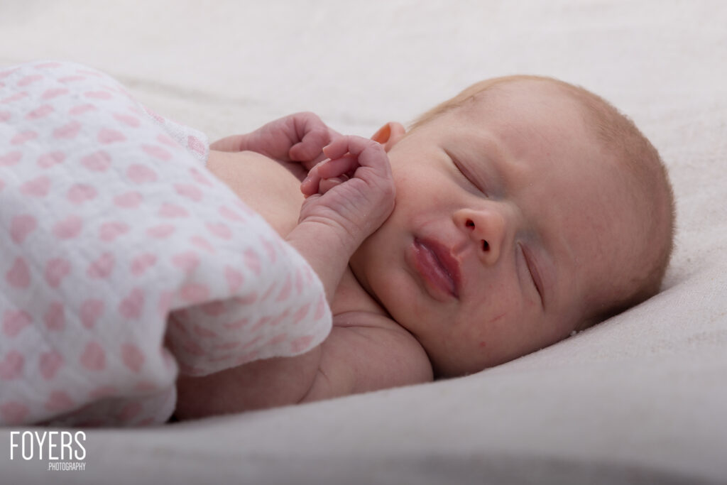 A newborn baby sleeps peacefully on white bedding, wearing a pink patterned garment with one hand visible near their face.