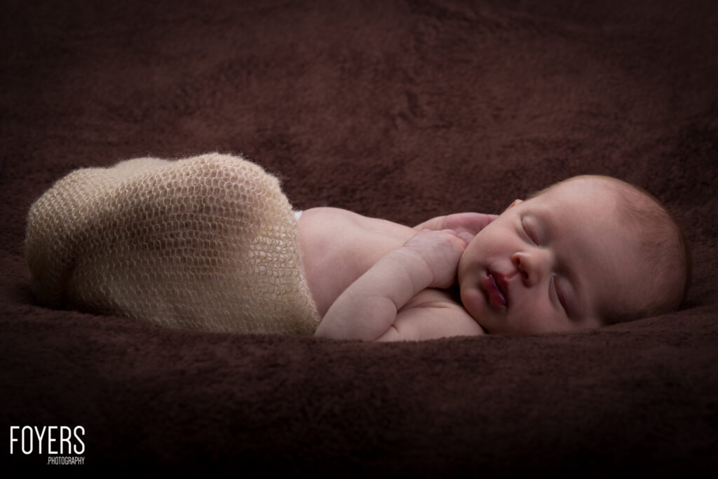 A newborn baby sleeps peacefully on a dark brown blanket, wrapped in a cream-coloured knitted wrap.