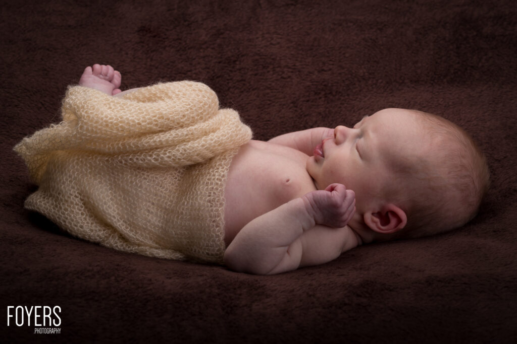 Newborn baby sleeping on a dark brown textured background, wrapped in a cream-coloured knitted blanket.