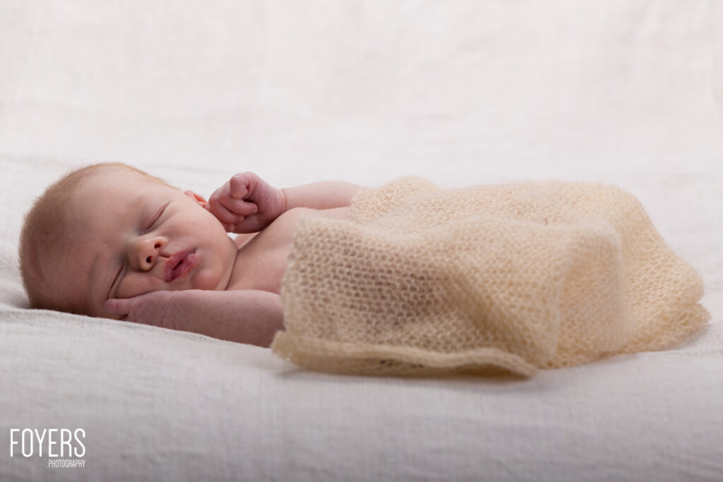 A newborn baby sleeps peacefully on a white surface, swaddled in a cream-coloured knitted blanket.