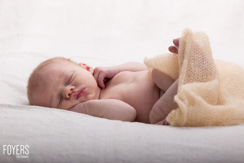 A newborn baby sleeps peacefully on white bedding, wrapped in a cream knitted blanket.